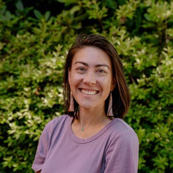 Hanako is wearing a short sleeved purple shirt and dangling earrings while smiling and standing in front of a green leafy background.